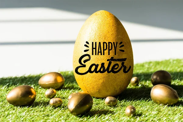 Большое страусиное яйцо со счастливой пасхальной надписью рядом с золотой курицей и перепелиными яйцами на зеленой траве — стоковое фото