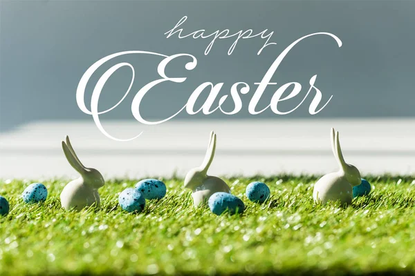 Conigli decorativi su erba verde vicino alle uova di quaglia blu con felice iscrizione pasquale — Foto stock