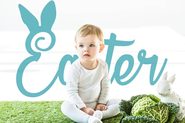 Criança sentada perto de couve lombarda e coelho decorativo na grama verde com letras azuis da Páscoa — Fotografia de Stock