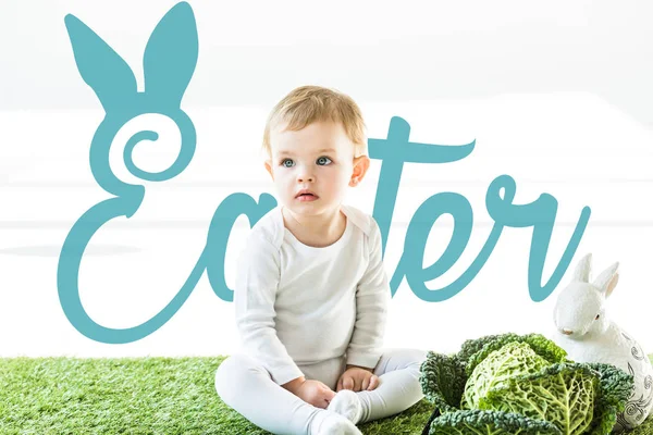Bambino seduto vicino al verza e coniglietto decorativo su erba verde con scritte pasquali blu — Foto stock