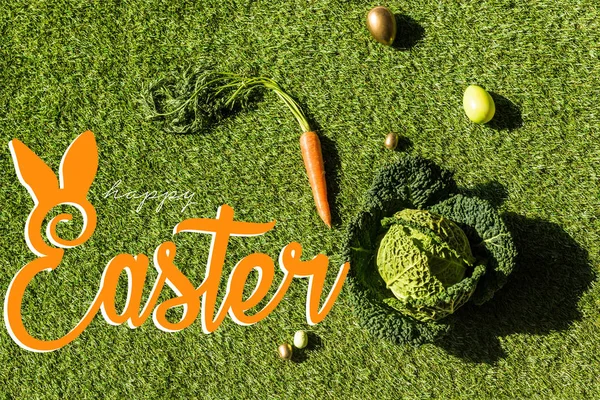 Top view of carrot, savoy cabbage and painted Easter eggs on green grass with happy Easter lettering — Stock Photo