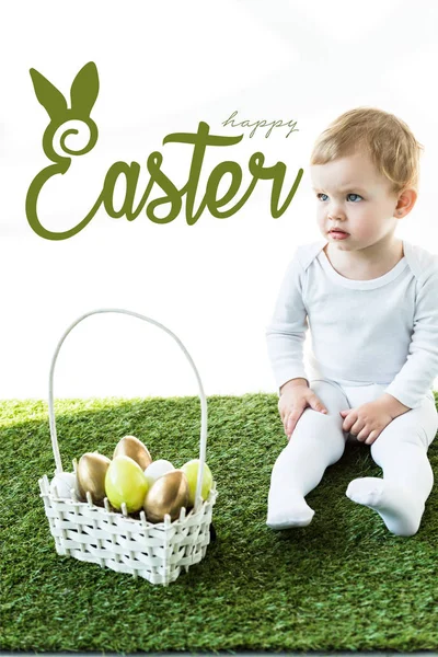 Child sitting on green grass near basket with Easter eggs and happy Easter lettering above — Stock Photo