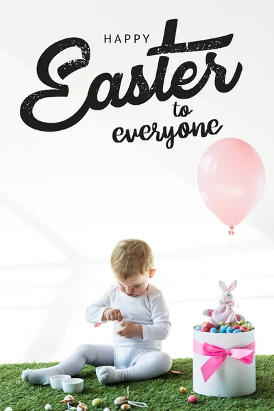 Carino bambino seduto vicino alla scatola con uova di quaglia colorate, coniglio giocattolo e palloncino su sfondo bianco con felice Pasqua a tutti lettering — Foto stock