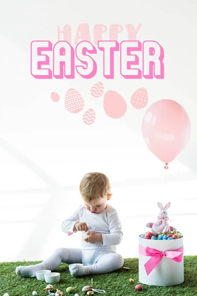 Cute baby sitting near box with colorful quail eggs, toy rabbit and air balloon on white background with happy Easter lettering — Stock Photo
