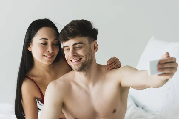 Handsome shirtless man making selfie with smartphone near attractive asian girlfriend — Stock Photo