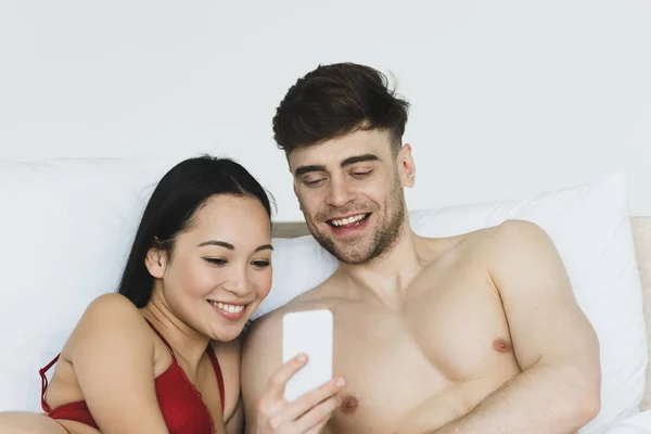 Sourire couple multiculturel à l'aide d'un smartphone tout en étant couché au lit — Photo de stock
