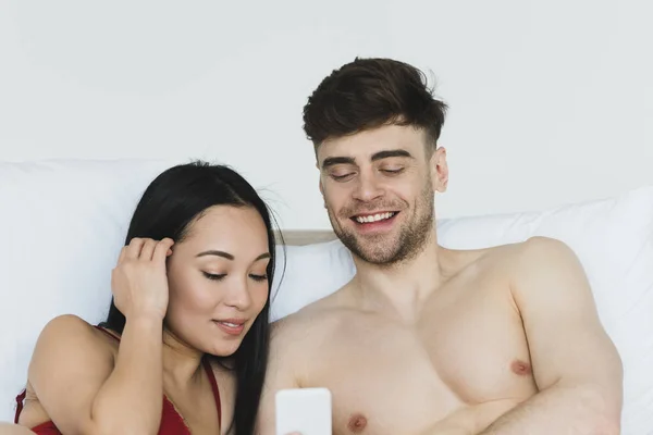 Jolie asiatique femme en rouge lingerie en utilisant smartphone avec torse nu petit ami au lit — Photo de stock