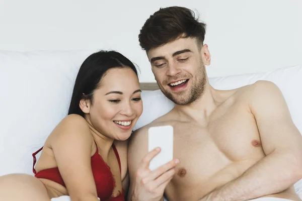 Belle femme asiatique en lingerie rouge en utilisant smartphone avec beau petit ami au lit — Photo de stock