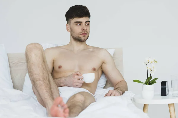 Selektiver Fokus eines gutaussehenden, nachdenklichen Mannes in Unterhosen, der mit einer Tasse Kaffee im Bett liegt — Stockfoto