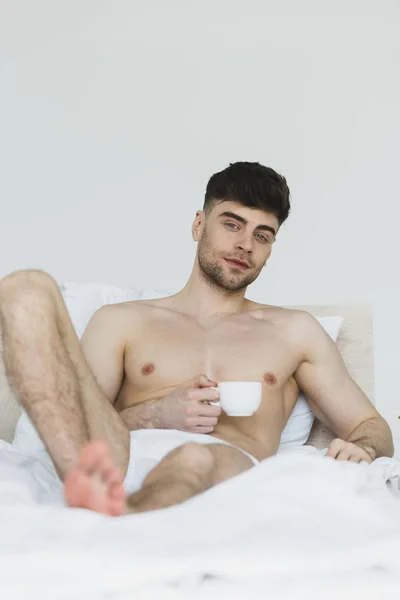 Вибірковий фокус доброзичливого молодого чоловіка в нижній білизні, лежить в ліжку з чашкою кави і дивиться на камеру — стокове фото