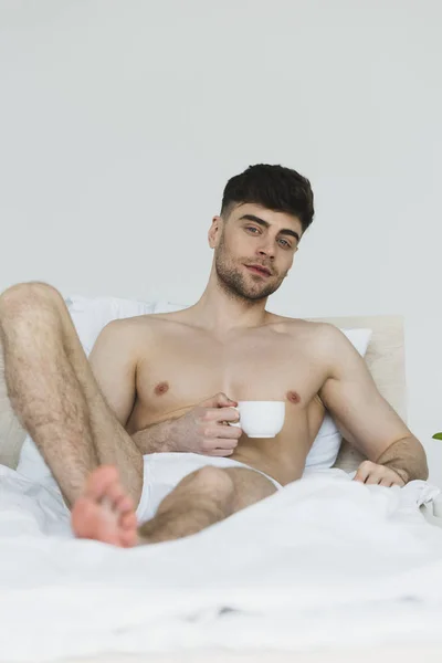 Selective focus of handsome smiling man in underwear lying in bed with coffee cup and looking at camera — Stock Photo