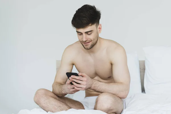 Красивий, усміхнений чоловік в нижній білизні сидить у ліжку і використовує смартфон — стокове фото