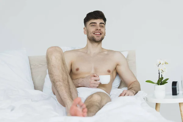 Selektiver Fokus eines gut aussehenden, lächelnden Mannes in Unterhosen, der mit einer Tasse Kaffee im Bett liegt — Stockfoto