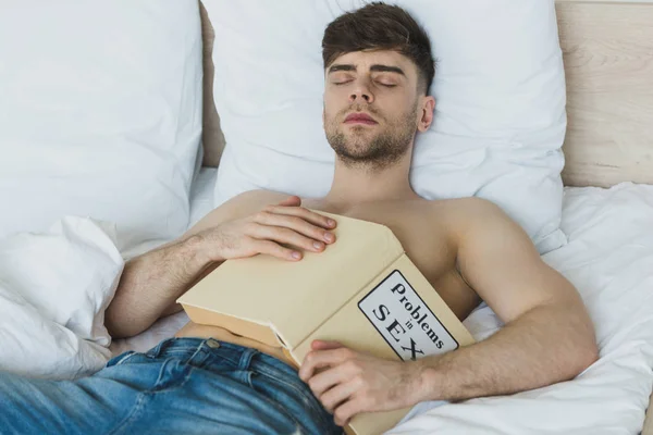 Guapo hombre sin camisa en pantalones vaqueros azules durmiendo con problemas en el libro de sexo - foto de stock