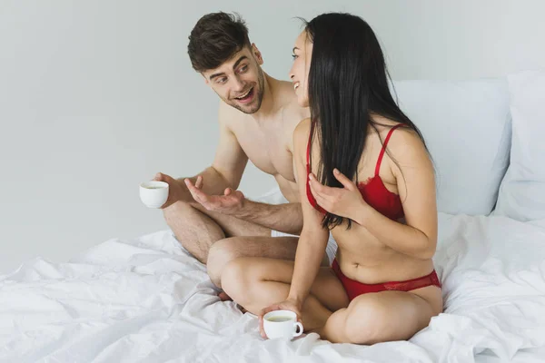 Cheerful multicultural couple talking while sitting on white bedding with coffee cups — Stock Photo