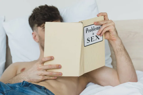 Selective focus of shirtless man in blue jeans reading problems in sex book while lying in bed — Stock Photo