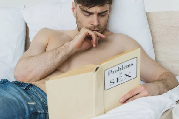 Selective focus of thoughtful shirtless man reading problems in sex book while lying in bed — Stock Photo