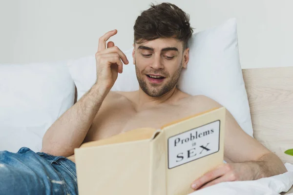 Selective focus of handsome shirtless man showing idea sign while reading problems in sex book — Stock Photo