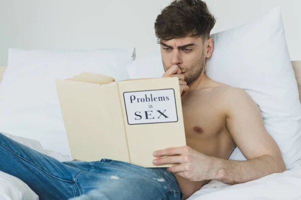 Handsome concentrated man in blue jeans reading problems in sex book while lying on white bedding — Stock Photo