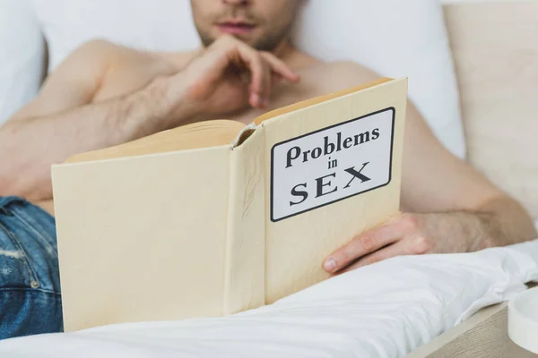 Teilansicht eines hemdlosen Mannes, der auf weißer Bettwäsche liegt und Probleme beim Lesen in einem Sexbuch hat — Stockfoto