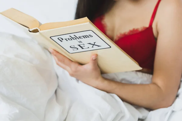 Vista parziale della donna in reggiseno rosso sdraiata a letto e problemi di lettura nel libro del sesso — Foto stock