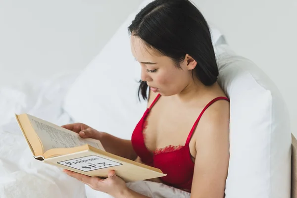 Hermosa, mujer asiática concentrada en sujetador rojo acostado en la cama y problemas de lectura en el libro de sexo - foto de stock