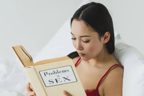 Selettivo fuoco di concentrato asiatico donna lettura problemi in sesso libro mentre sdraiato in letto — Foto stock