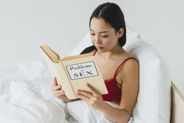 Beautiful asian woman in red bra reading problems in sex book while lying in bed — Stock Photo