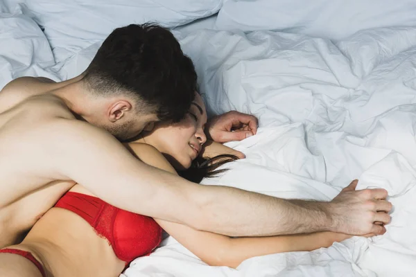 Shirtless man kissing beautiful asian girlfriend in red lingerie while lying on white bedding — Stock Photo