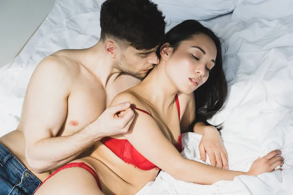 Handsome shirtless man kissing passionate asian girlfriend in red lingerie — Stock Photo