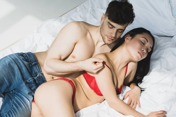 Sexy shirtless man kissing passionate asian girlfriend in red lingerie while lying on white bedding — Stock Photo