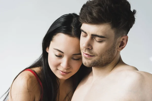 Beautiful, pensive asian woman hugging handsome shirtless boyfriend with closed eyes on grey — Stock Photo