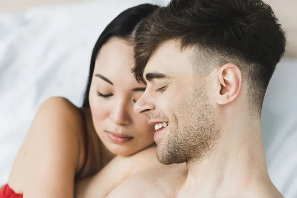 Happy multicultural couple hugging while lying on white bedding together — Stock Photo