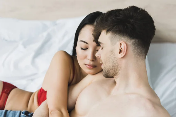 Bela ásia mulher no vermelho lingerie abraçando sem camisa namorado enquanto deitado no cama juntos — Fotografia de Stock