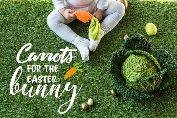 Partial view of child holding toy carrot while sitting near Easter eggs and savoy cabbage on green grass with carrots for the Easter bunny lettering — Stock Photo
