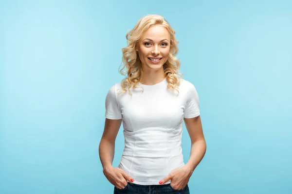 Fröhliche blonde Frau blickt in die Kamera, während sie mit den Händen in den Taschen auf blau steht — Stockfoto