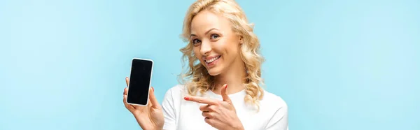 Панорамний знімок весела блондинка, що вказує пальцем на смартфон з порожнім екраном ізольовано на синьому — стокове фото
