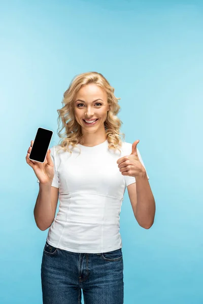 Mujer rubia alegre mostrando el pulgar hacia arriba mientras sostiene el teléfono inteligente con pantalla en blanco en azul - foto de stock