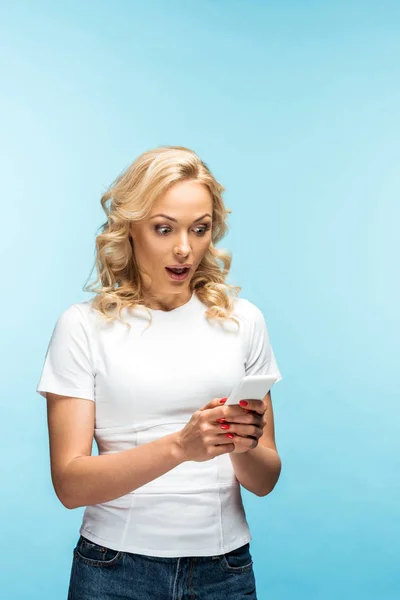 Femme blonde choquée regardant smartphone et debout sur bleu — Photo de stock