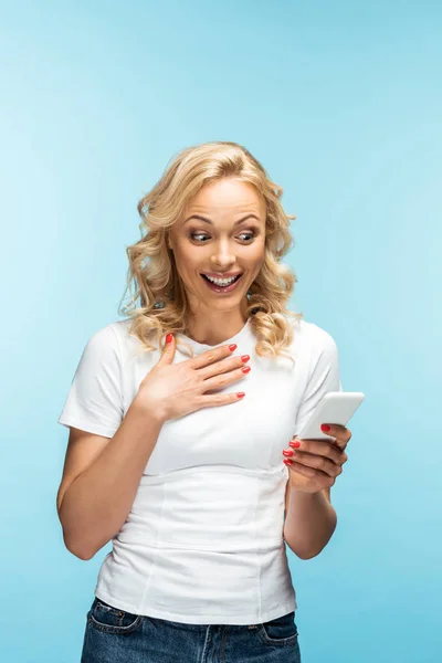 Щаслива блондинка дивиться на смартфон, стоячи на синьому — стокове фото