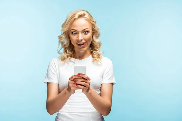 Счастливая блондинка, демонстрирующая язык, глядя на смартфон на голубом — стоковое фото