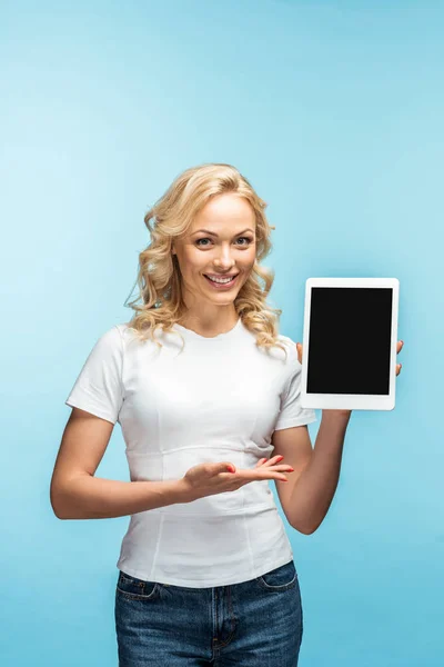 Mujer rubia feliz gesto mientras sostiene la tableta digital con pantalla en blanco en azul - foto de stock