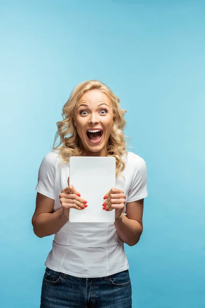 Mujer rubia impactada mirando a la cámara mientras sostiene la tableta digital en azul - foto de stock