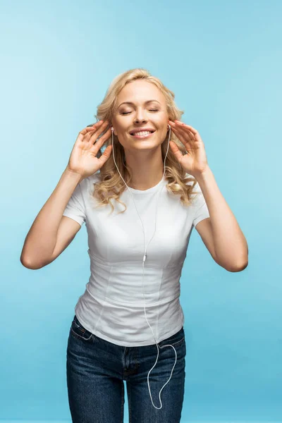 Cheerful woman with closed eyes listening music in earphones on blue — Stock Photo