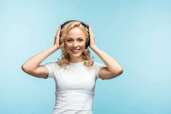 Happy blonde woman listening music and touching black headphones on blue — Stock Photo