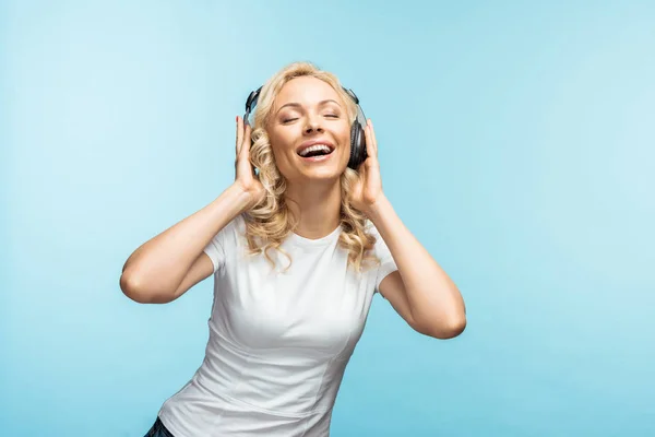 Femme blonde heureuse avec les yeux fermés écouter de la musique dans des écouteurs noirs sur bleu — Photo de stock