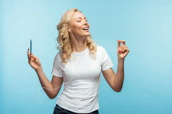 Happy woman with closed eyes listening music in earphones while holding smartphone on blue — Stock Photo