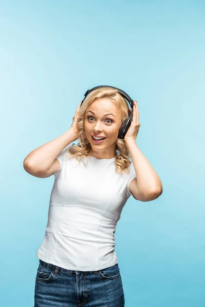 Surprised blonde woman listening music in black headphones on blue — Stock Photo