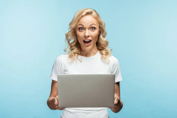 Surprised blonde woman holding laptop and looking at camera on blue — Stock Photo