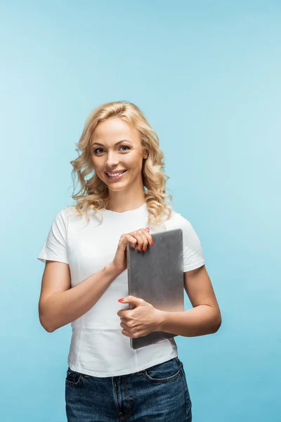 Glückliche blonde Frau hält Laptop in der Hand und schaut in die Kamera auf blau — Stockfoto